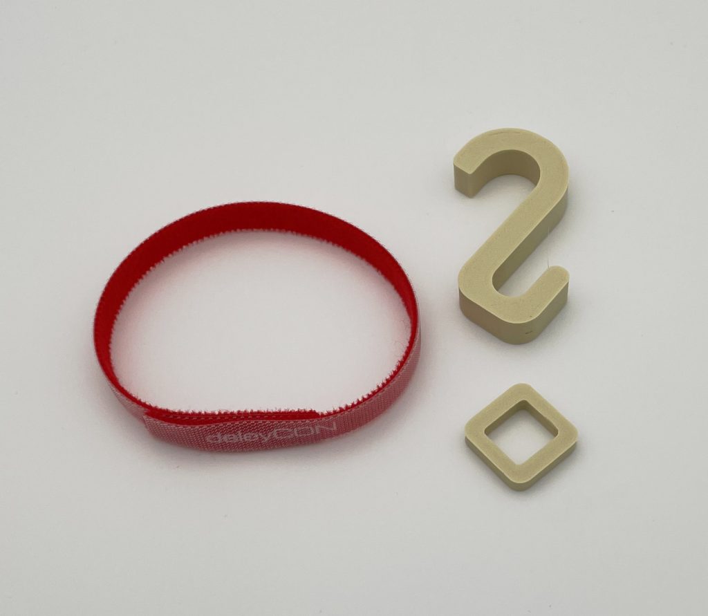 Die Einzelteile, die im Produkt "Schranksicherung für Hängeschränke" für Eriba Troll oder Touring entwickelt wurde. Das Produkt besteht aus einem Klettband, einem Haken und einer Öse. 